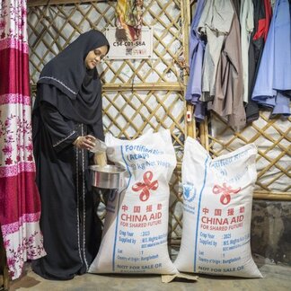 A person stands inside a bamboo-walled shelter and pours grain from a container into a cooking pot. Two large sacks of food assistance marked with WFP and donor branding are positioned nearby, illustrating household food support and access to staple commodities.
