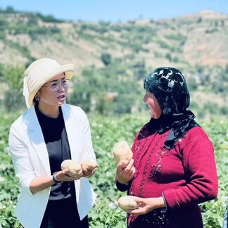 Two people stand in a cultivated field holding freshly harvested potatoes and talking with one another. Green crops stretch across the foreground, while rolling hills and agricultural land form the backdrop, illustrating local farming and food production activities.