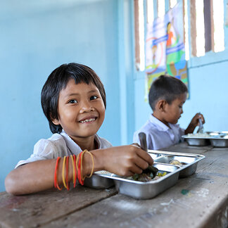 A student sits at a wooden classroom desk eating a meal from a metal tray. Another student is seated in the background with a similar tray. The classroom has light blue walls, and a colorful poster is visible near the windows.