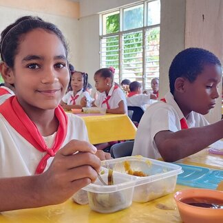 Children in uniform eat meals from plastic containers at a yellow-covered table inside a bright classroom with large windows.