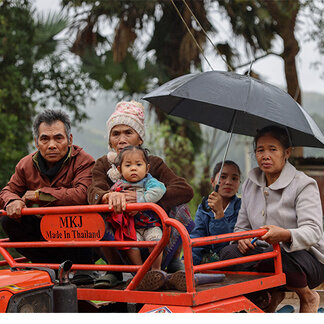 Several community members sit on a small agricultural vehicle during rainy weather. One person holds an umbrella while others sit nearby, including a young child. Trees and vegetation are visible in the background, and the scene depicts transportation in a rural area following adverse weather conditions.