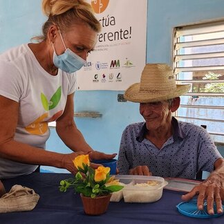 A person serves food to another seated individual at a table with a flower pot, under a banner promoting sustainable food systems.