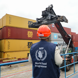 A supply chain officer wearing a hard hat and WFP vest stands facing a crane between shipping containers