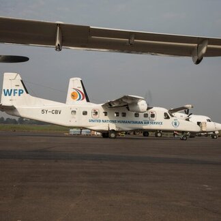 White aircraft labelled 'UNITED NATIONS HUMANITARIAN AIR SERVICE' and 'WFP', with personnel and other planes visible in the background.