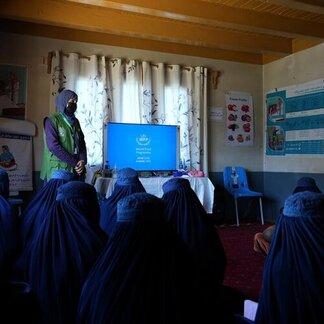 The backs of women wearing burqas and sat facing towards a training session. The instructor is wearing a green gilet and is on the right of a TV screen showing the WFP logo.