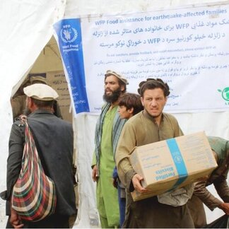 People receive WFP aid in a tent bearing a multilingual banner. One person holds a box labelled "Fortified Biscuits for earthquake victims."