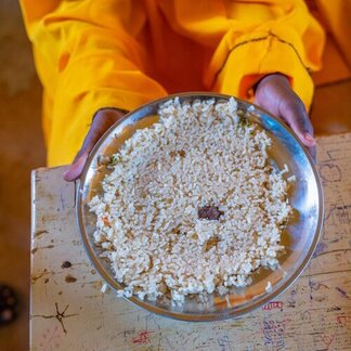 A child holds a metal plate filled with a small serving of rice, showing the limited meal available in a school setting.