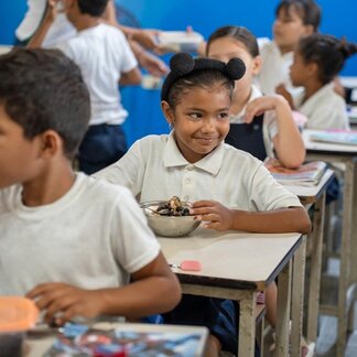 Children sit at classroom desks eating meals from metal bowls, with notebooks and lunch containers nearby. Others are visible in the background near a blue wall. Photo: WFP/Gustavo Vera