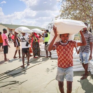 People carrying large white sacks of supplies in an outdoor distribution area on a sunny day.