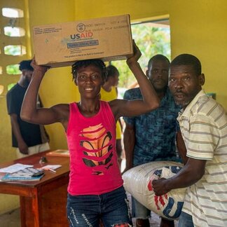 Person in a pink shirt holds a large cardboard box above their head while others sit and stand around a table in a bright yellow room.