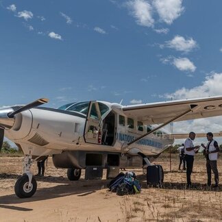 Small aircraft on a dirt airstrip with luggage and people standing nearby under a clear blue sky.