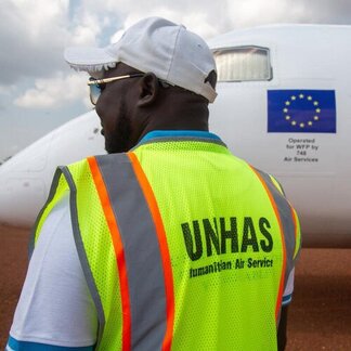 UNHAS staff member in a high-visibility vest stands beside a humanitarian aircraft on a runway, supporting air transport operations.