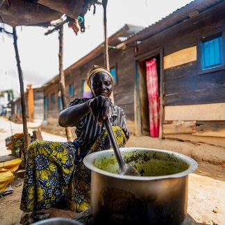 Person cooks food in a large pot outside a wooden home in a settlement, preparing a meal over an open fire in daylight.