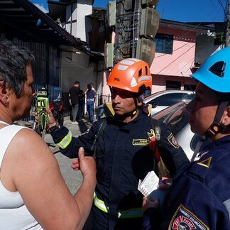 Helmeted emergency responders speak with a resident beside damaged buildings and overhead utilities on a street in Colombia.