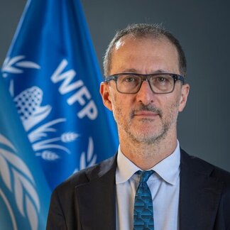 Portrait of a WFP official standing in front of WFP and United Nations flags, wearing a dark suit, white shirt and blue patterned tie.