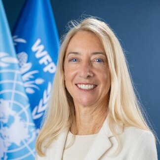 Person with long blonde hair stands before two blue flags with white WFP logos; face is blurred for privacy.