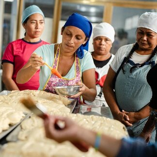 People in aprons and head coverings prepare bread dough on trays, brushing glaze from bowls in a bright kitchen during a group baking activity. Photo: WFP/Gustavo Vera.