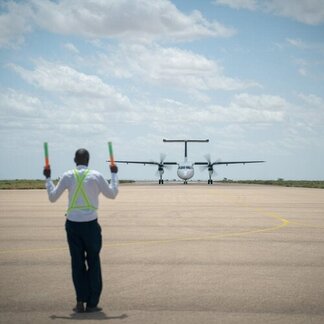 An airport worker guides an incoming propeller aircraft on the runway, raising signal batons as the plane approaches under a bright sky.