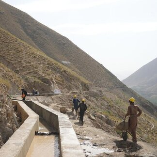 Men in yellow hats, some with wheelbarrows, working on an irrigation canal on a mountainside. 