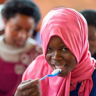 A girl in a pink headscarf eats a spoonful of food in a classroom.