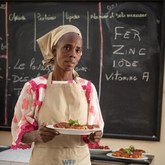 WFP-supported nutrition training participant wearing an apron presents a prepared meal in a classroom, with nutrition information on a chalkboard behind.