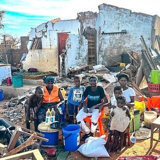 A group of seven adults and children are standing and sitting in front of a ruined building.