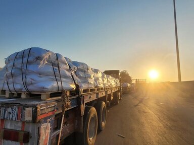 A truck loaded with large white bags secured with black straps is parked on a road at sunset, casting long shadows and creating a glow.